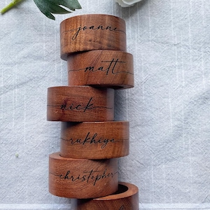 May include: Six wooden napkin rings with names engraved on them. The names are Joanne, Matt, Nick, Rukhiya, Christopher, and Carly. The rings are stacked on top of each other on a white linen tablecloth.