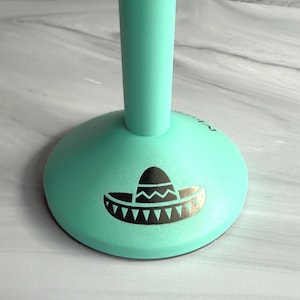May include: A mint green stand with a circular base featuring a black graphic print of a sombrero. The object has a smooth matte finish and a vertical cylindrical stem extending upward from the center of the rounded base.