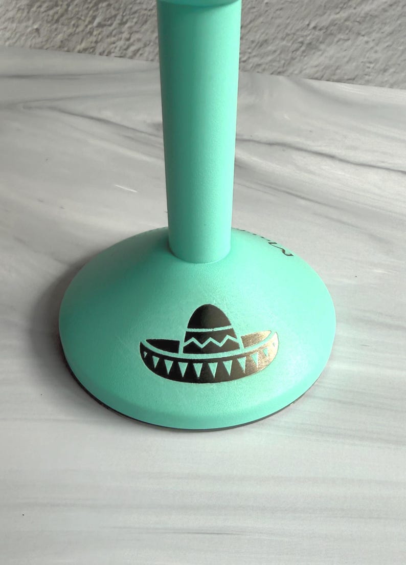 May include: A mint green stand with a circular base featuring a black graphic print of a sombrero. The object has a smooth matte finish and a vertical cylindrical stem extending upward from the center of the rounded base.