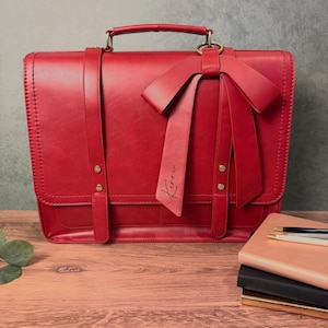 May include: A red leather briefcase with a bow detail. The briefcase has a top handle, a shoulder strap, and a flap closure. The bow has the name "Kerri" written on it. The briefcase is on a wooden surface.