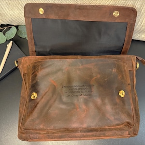 Pu&ograve; includere: Una borsa a tracolla in pelle marrone con chiusura a patta fissata da due bottoni a pressione in metallo dorato. La tasca frontale presenta una citazione incisa: The foundation of your life and its strength is completely up to you. -Dr. Corey M. Shores.