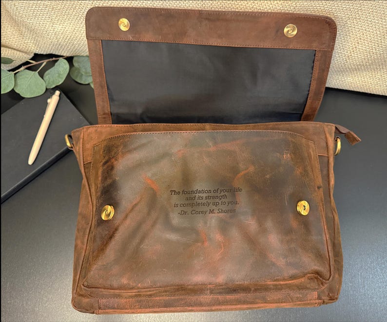 Pu&ograve; includere: Una borsa a tracolla in pelle marrone con chiusura a patta fissata da due bottoni a pressione in metallo dorato. La tasca frontale presenta una citazione incisa: The foundation of your life and its strength is completely up to you. -Dr. Corey M. Shores.
