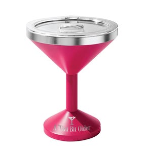 May include: A hot pink martini glass-shaped tumbler with a silver metallic rim and a clear plastic lid. The base features a white graphic of a martini glass and the text A 'Tini Bit Older printed in a clean, sans-serif font on the front.