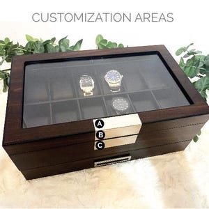 Puede incluir: Una caja para relojes de madera marr&oacute;n oscuro con tapa de cristal y diez compartimentos interiores. El frente presenta dos placas met&aacute;licas plateadas etiquetadas A, B y C para &aacute;reas de personalizaci&oacute;n y un caj&oacute;n inferior con asa.