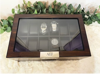 May include: A rectangular dark wood watch storage box with a glass lid and ten interior compartments. Three wristwatches are displayed inside on black cushions. A silver-toned metal plate on the front is engraved with the letters AEF.