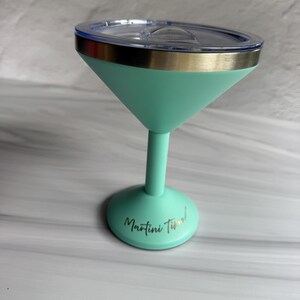 May include: A mint green insulated martini glass with a metallic silver rim and a clear plastic lid. The base of the glass features the text Martini Time! in a cursive black font. The stem and base are a solid mint green color.