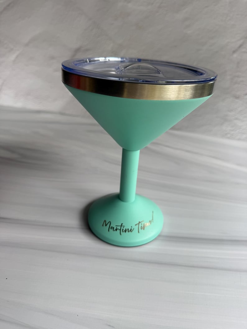 May include: A mint green insulated martini glass with a metallic silver rim and a clear plastic lid. The base of the glass features the text Martini Time! in a cursive black font. The stem and base are a solid mint green color.