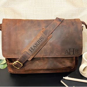 May include: A brown leather messenger bag with a flap closure and adjustable shoulder strap. The strap has the name "HARRIS" engraved on it. The bag also features the initials "AHB" and a gold-colored buckle.