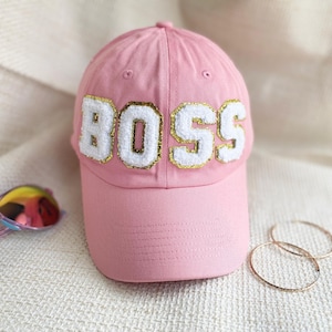 May include: Pink baseball cap with a white "BOSS" embroidered on the front. The cap has a gold outline around the letters.