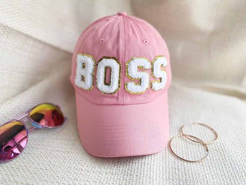 May include: Pink baseball cap with a white "BOSS" embroidered on the front. The cap has a gold outline around the letters.