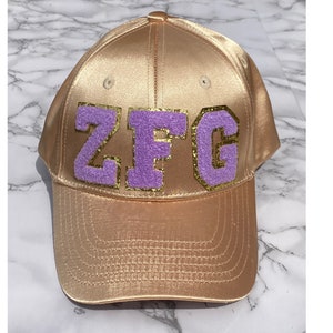 May include: A gold satin baseball cap with a purple felt patch that says "ZFG" in gold glitter letters.