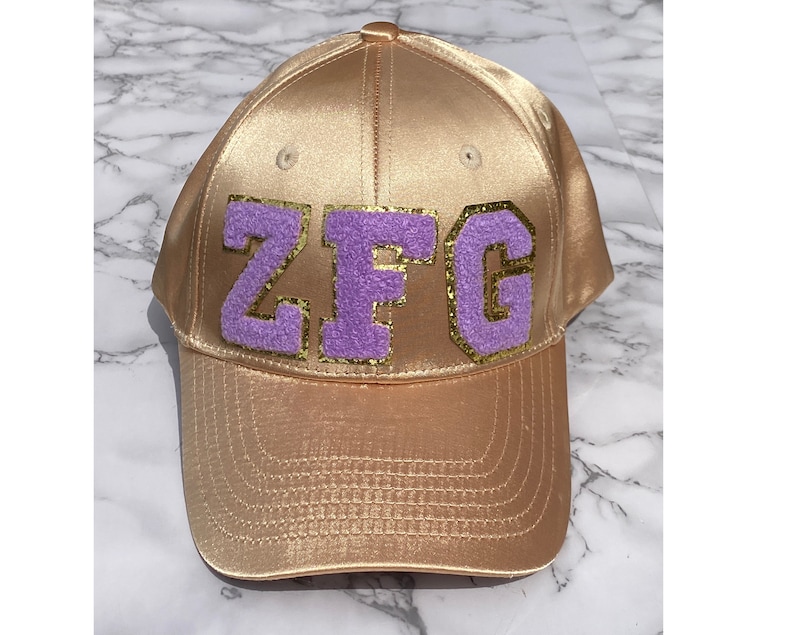 May include: A gold satin baseball cap with a purple felt patch that says "ZFG" in gold glitter letters.