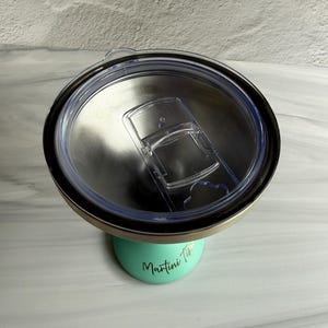 May include: A mint green stainless steel tumbler with a clear plastic lid featuring a sliding closure. The tumbler has the text "Martini Time" printed in a black cursive font on the side. The interior of the tumbler is metallic silver.
