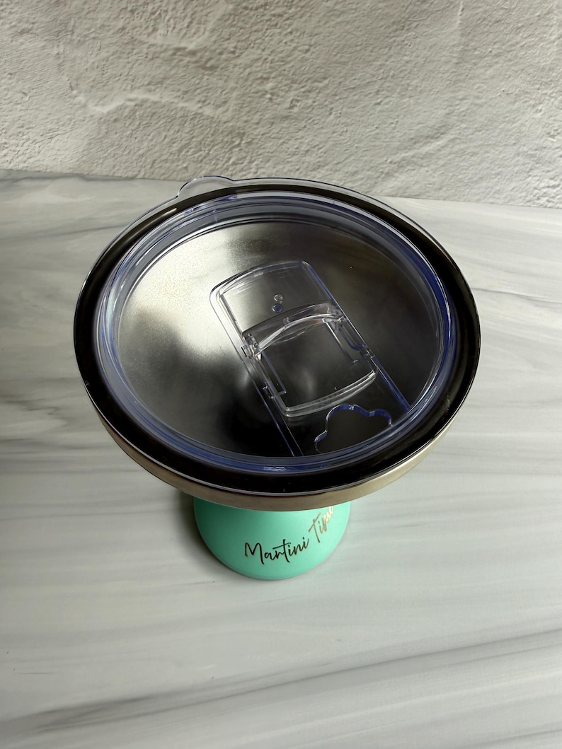 May include: A mint green stainless steel tumbler with a clear plastic lid featuring a sliding closure. The tumbler has the text "Martini Time" printed in a black cursive font on the side. The interior of the tumbler is metallic silver.