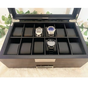 Walnut finished wood watch display case with black faux leather interior and watch pillows. Watch box case is displayed with glass lid open.