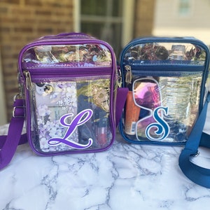 May include: Two clear plastic crossbody bags with purple and blue trim. The bag on the left has a purple strap and the letter "L" on the front. The bag on the right has a blue strap and the letter "S" on the front.