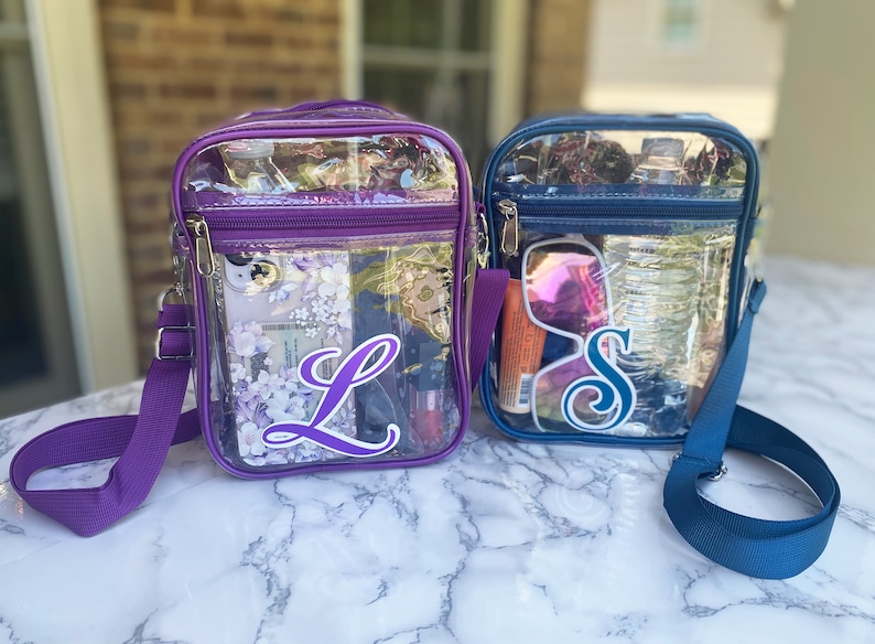 May include: Two clear plastic crossbody bags with purple and blue trim. The bag on the left has a purple strap and the letter "L" on the front. The bag on the right has a blue strap and the letter "S" on the front.