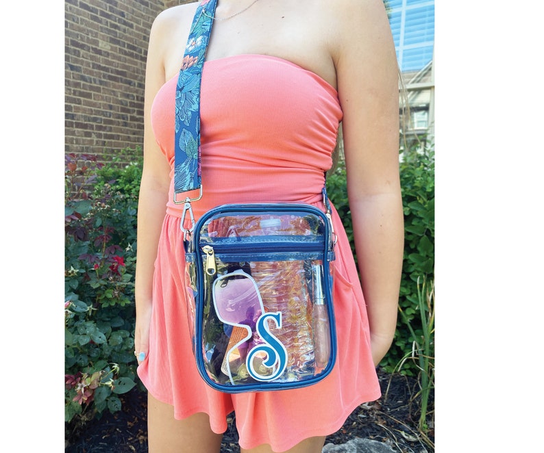 May include: A clear plastic crossbody bag with a navy blue trim and a floral patterned strap. The bag has a zipper closure and a clear pocket on the front. The bag is filled with various items, including sunglasses, a phone, and a water bottle. The bag has a blue and white monogrammed letter "S" on the front.