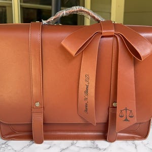 May include: A cognac-colored leather briefcase featuring a top handle wrapped in protective plastic, two vertical straps with gold-tone metal studs, and a large decorative bow. One ribbon of the bow displays the text Sharice N. Bland, J.D.
