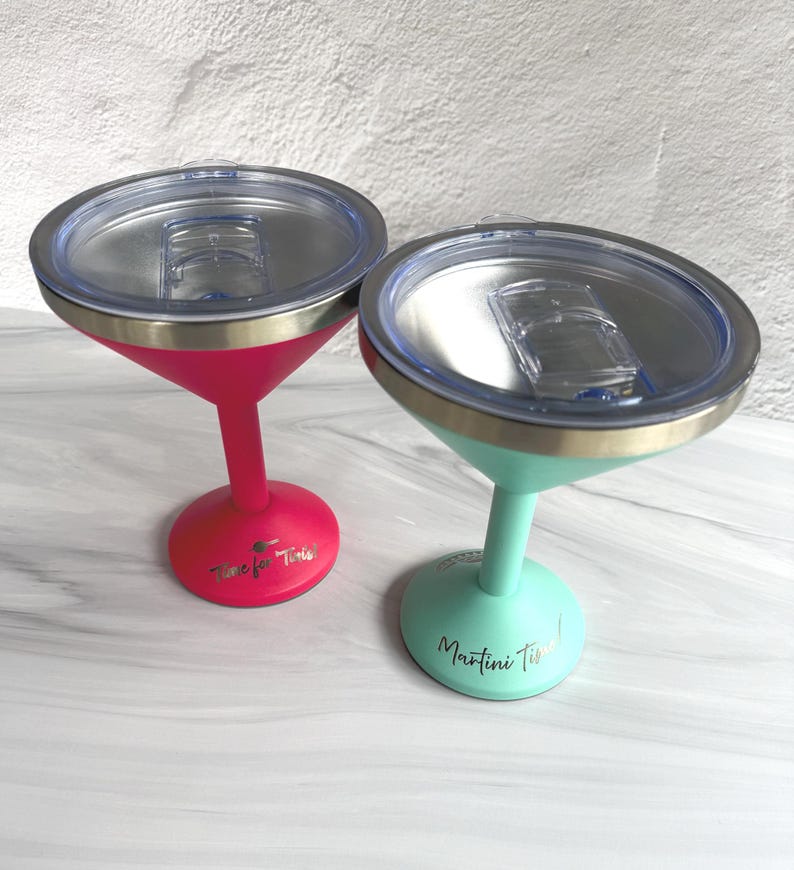 May include: Two stainless steel martini glasses with clear plastic lids. One glass is hot pink with the text Time for Tinis! and the other is mint green with the text Martini Time! printed in gold on the base of each stem.