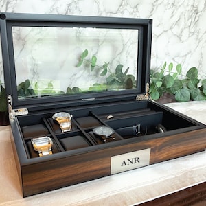 May include: A dark wood watch box with a glass top and a silver plaque with the initials "ANR" engraved on it. The box has multiple compartments for storing watches and other accessories.