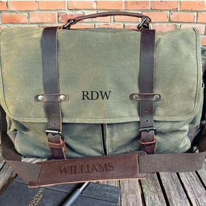 May include: A green canvas messenger bag with brown leather straps and accents. The bag features the initials "RDW" and the name "WILLIAMS" engraved on the leather. It has a top handle and a shoulder strap.