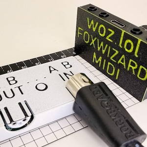May include: A black and white electronic MIDI interface device with the text WOZ.LOL FOXWIZARD MIDI printed in yellow. The device features a white panel labeled A B OUT and A B IN, a black cable connector, and is positioned on a grid measuring tool.