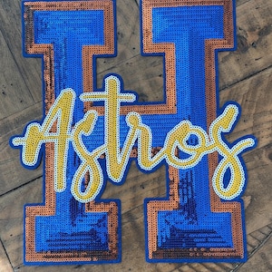 May include: A blue and orange sequined patch in the shape of the letter "H" with the word "Astros" in a yellow script font. The patch is likely for a sports team, possibly baseball.