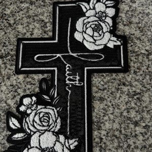 May include: Black cross-shaped patch with white floral accents and the word "Faith" in script. The patch features a detailed design with embroidered roses and leaves, suitable for clothing or accessories.