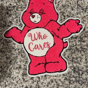 May include: A pink Care Bear-style rug with a white border. The bear has a sad expression and the words "Who Cares" embroidered on its belly. The rug is on a speckled gray surface.