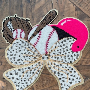 May include: A baseball-themed decorative piece featuring a baseball, bat, glove, and helmet. The design includes a large bow with a sequined leopard print and gold trim. The baseball has red stitching, and the helmet is bright pink.