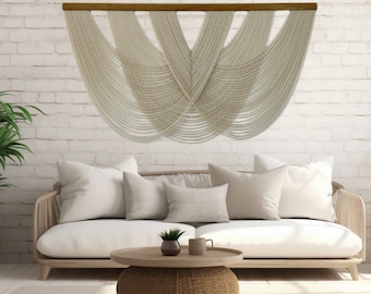 May include: A white macrame wall hanging with a wooden dowel rod at the top. The macrame is in a geometric pattern with a V-shape in the center. The wall hanging is hanging on a white brick wall above a white couch with pillows and a wooden coffee table.