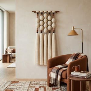 May include: A cream-coloured fibre art wall hanging with a wooden top and bottom. The design features multiple rows of round, off-white spheres and long, cream-coloured tassels. The wall hanging is displayed above a brown armchair with a plaid throw.