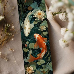 Puede incluir: Un marcapáginas con una ilustración detallada de dos peces koi nadando en un estanque con nenúfares. Un pez es blanco con manchas rojas, el otro es naranja y blanco. El fondo es verde oscuro.