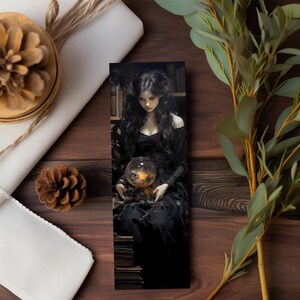 May include: A bookmark featuring a gothic illustration of a woman in a black dress holding a glowing glass orb with a skull inside. The woman has long dark hair and is seated in a library setting. The bookmark is rectangular and vertical.