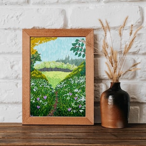 May include: A framed landscape painting depicts a vibrant green meadow with a path leading to a distant forest. The artwork features a blue sky, lush foliage, and purple flowers. A dark brown vase with dried wheat stalks sits to the right.