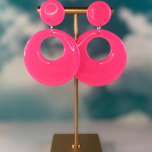 Puede incluir: Un par de pendientes de aro rosa con un dise&ntilde;o grande, redondo y abierto. Los pendientes se muestran en un soporte dorado.