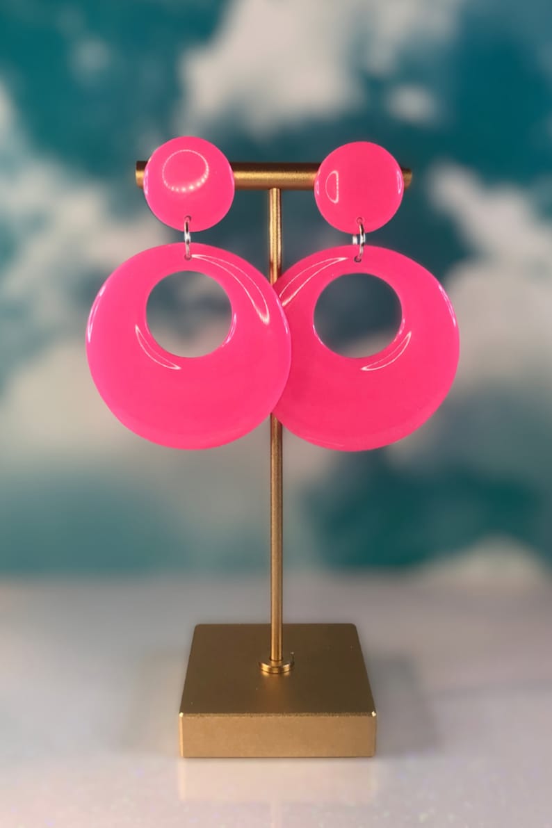 Puede incluir: Un par de pendientes de aro rosa con un dise&ntilde;o grande, redondo y abierto. Los pendientes se muestran en un soporte dorado.