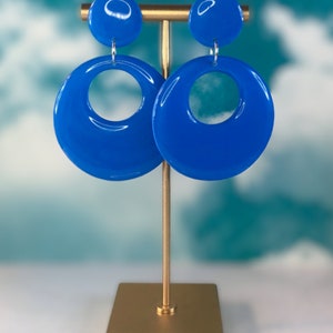 Puede incluir: Un par de pendientes de aro azules con un dise&ntilde;o de c&iacute;rculo grande. Los pendientes est&aacute;n en un soporte dorado sobre un fondo azul y blanco.