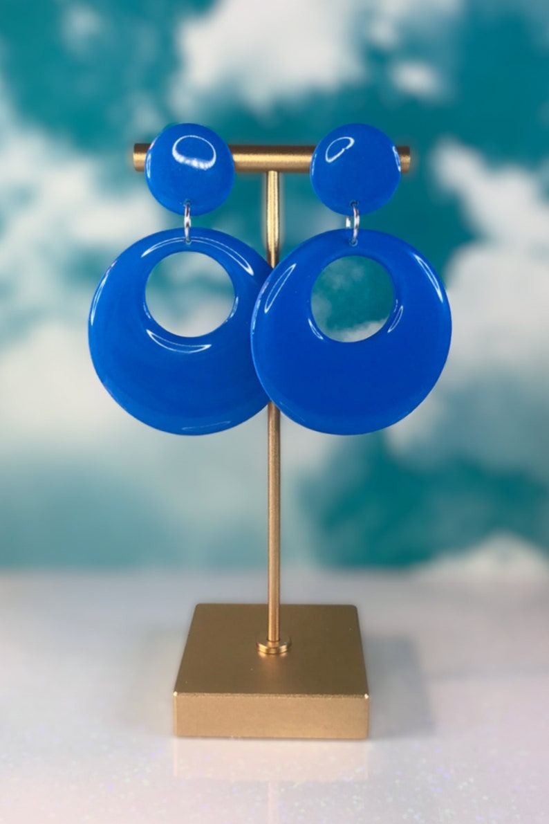Puede incluir: Un par de pendientes de aro azules con un dise&ntilde;o de c&iacute;rculo grande. Los pendientes est&aacute;n en un soporte dorado sobre un fondo azul y blanco.
