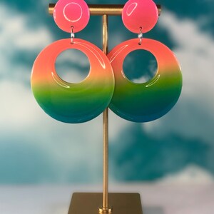 Puede incluir: Un par de pendientes de aro degradados con un degradado de rosa, naranja, amarillo, verde y azul. Los pendientes est&aacute;n colgados de un soporte dorado.