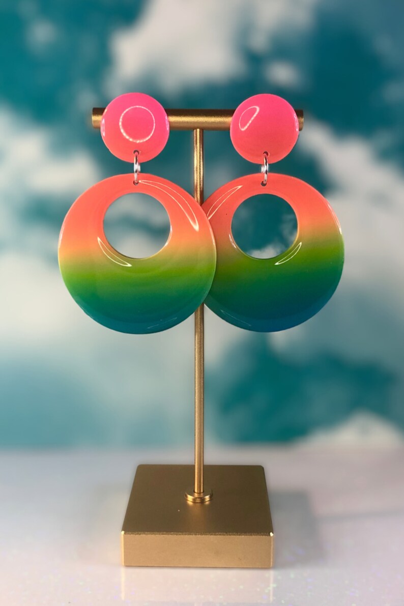 Puede incluir: Un par de pendientes de aro degradados con un degradado de rosa, naranja, amarillo, verde y azul. Los pendientes est&aacute;n colgados de un soporte dorado.