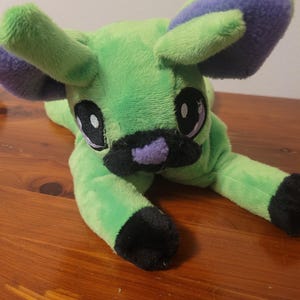 May include: A plush toy in the shape of a stylized animal, primarily green with purple accents on the ears and nose. The toy has large, round eyes and black paws. The plush is lying down on a wooden surface.