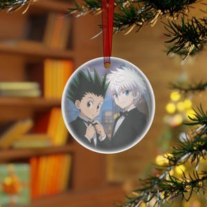 May include: A clear, round Christmas ornament featuring two anime characters in formal attire. One character has green spiky hair, the other has white hair. The ornament hangs from a red ribbon, with a blurred background of a Christmas tree and a bookshelf.