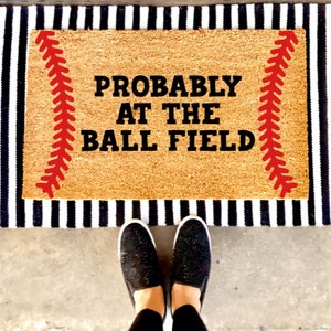 May include: A brown coir doormat with black and white stripes around the edges. The doormat has red stitching on either side and the text "PROBABLY AT THE BALL FIELD" in black.