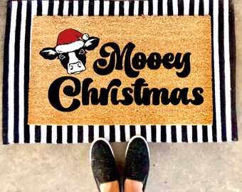 May include: A rectangular tan coir doormat layered over a black and white striped rug. The doormat features a black and white cow illustration wearing a red Santa hat and the black text "Mooey Christmas" in a bold, cursive, retro-style font.