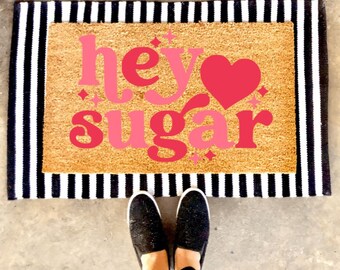 May include: A pink and white doormat with the text "hey sugar" and a pink heart. The doormat has a black and white striped border.