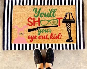 May include: A brown coir doormat with black and white stripes around the edges. The doormat has a white background with the text "You'll SHOOT your eye out, kid!" in green, red, and black. There is a pink bunny rabbit with glasses and a black lamp with a leg in the shape of a woman's leg.
