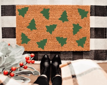 May include: A rectangular doormat with a brown coir surface features a pattern of green Christmas trees. The mat is placed on a black and white checkered rug. Black boots are visible in the foreground.