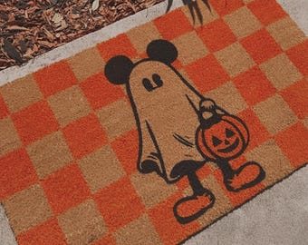 May include: A brown and orange checkered doormat with a black and white illustration of a ghost wearing a Mickey Mouse hat and holding a jack-o-lantern.
